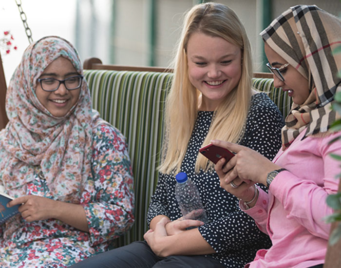Three diverse female students smiling and looking at a phone together on a bench