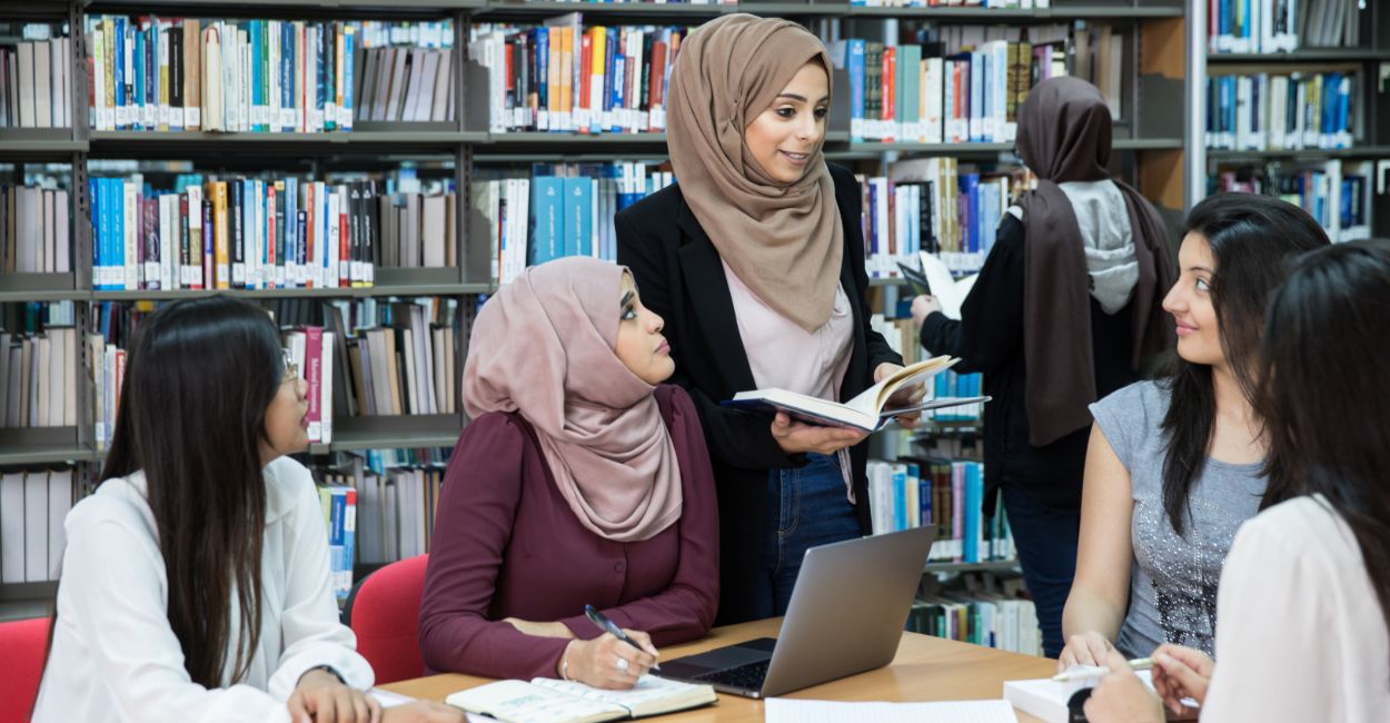 Group of diverse female students studying and discussing at a library table with books and laptops.