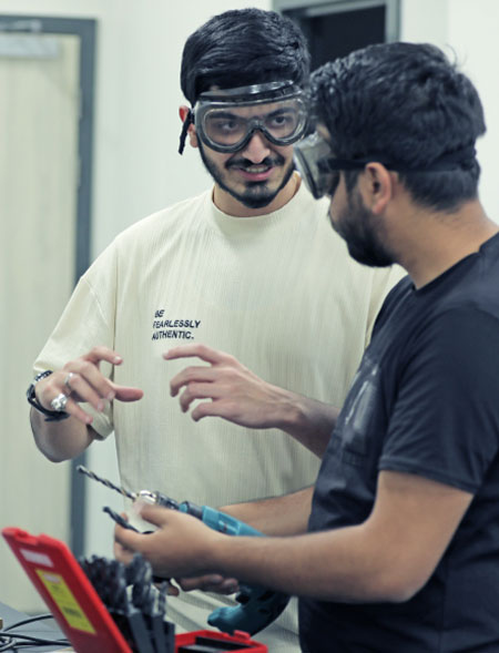 Two students wearing safety goggles discussing drill tools in a lab setting.