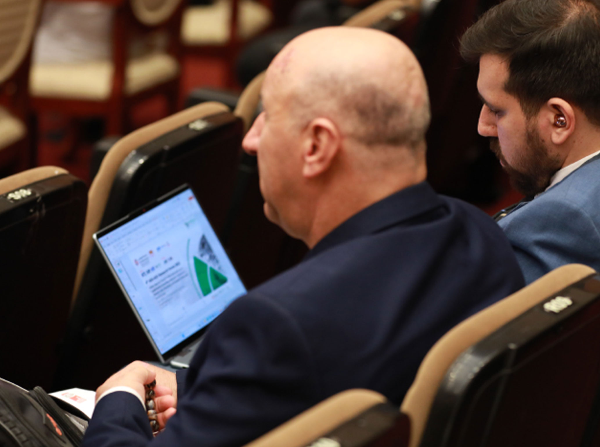 Two men seated in an auditorium reviewing a presentation slide on a laptop during a conference.