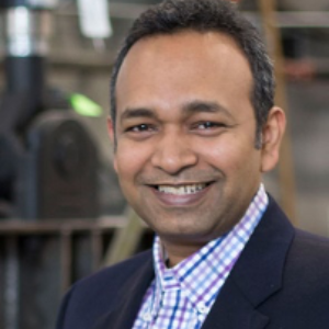 Smiling man in blazer and checkered shirt standing in an industrial or workshop setting.